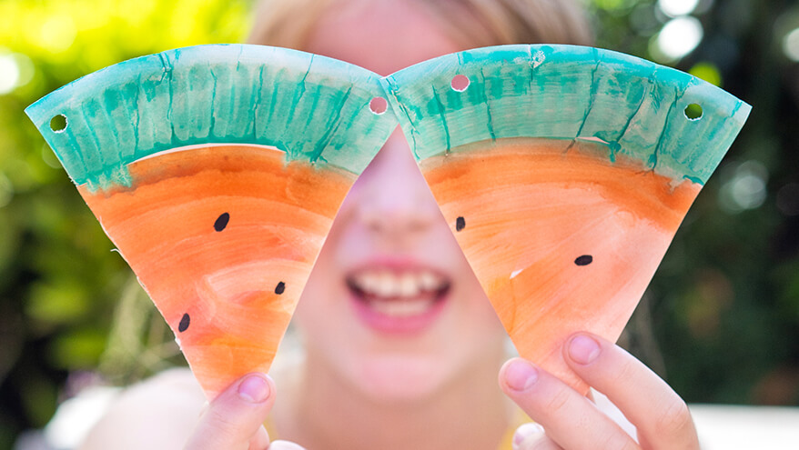 Ein Mädchen im Grundschulalter hält zwei Wassermelonen-Dreiecke aus Papptellern von ihr Gesicht und lacht dabei. Im Hintergrund ist das verschwommene Grün eines Gartens sichtbar. Foto: Johanna Rundel 